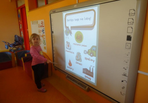 Dziewczynka stoi pod tablicą interaktywną i wskaźnikiem pokazuje pożar.